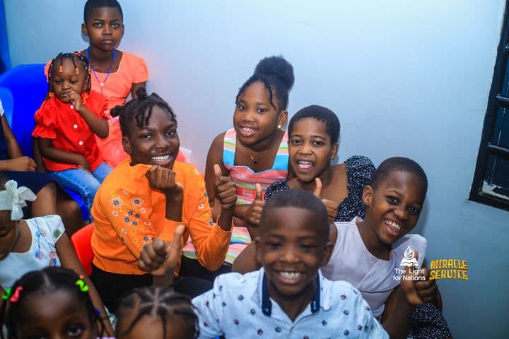 Five of the older children crowded together, grinning and giving thumbs-up, with a younger girl in red seated behind them.