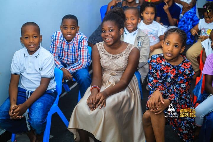 Four children seated in a row against a pale wall, calm and looking towards the camera, a girl in a gold dress in the middle.