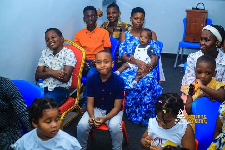 Older children at the back of the room, one of them holding a toddler on her lap, with younger children seated in front of them.